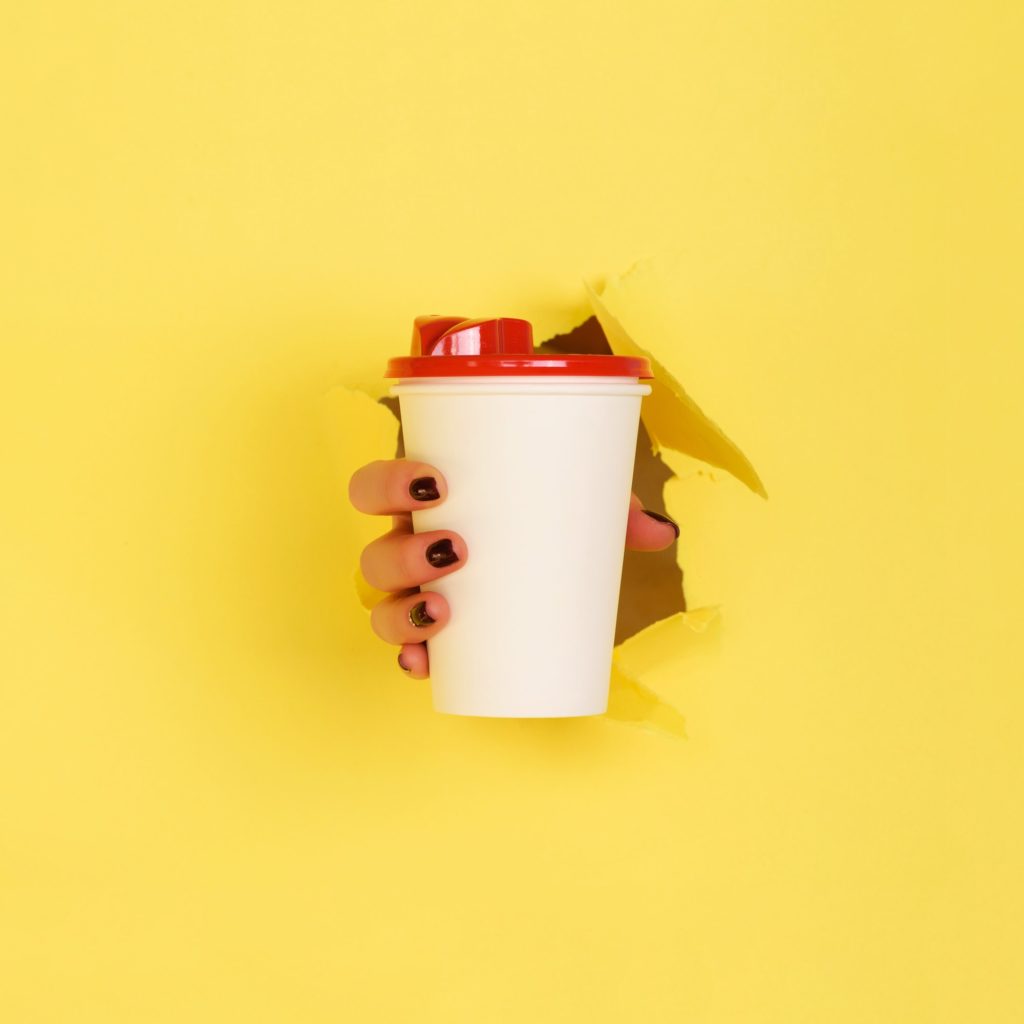 Female hand holding white paper mug on yellow background. Take away coffee cup concept. Mock up with